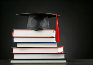A stack of books is topped with a black graduation cap featuring a red tassel, set against a dark background.