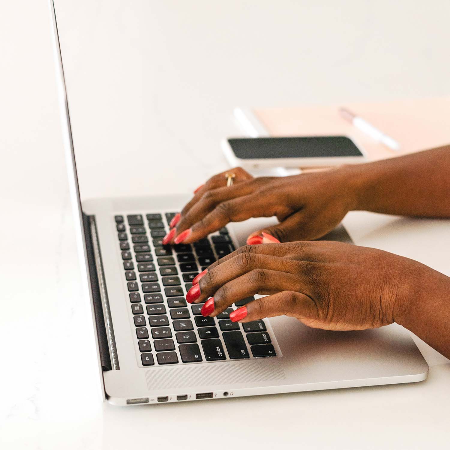 A person with red-painted nails types on a silver laptop keyboard. A smartphone, notebook, and pen are placed on the white desk beside the laptop.