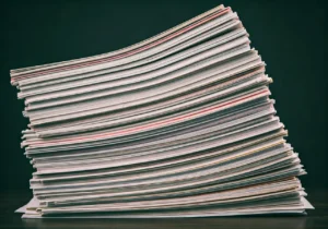 A large stack of magazines or documents is neatly piled on a dark surface, viewed from the side. The papers are slightly curved, displaying colorful edges against a dark background.