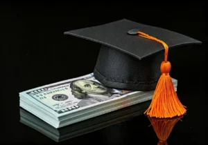 A graduation cap with an orange tassel rests on a stack of U.S. hundred dollar bills, symbolizing the cost or value of education, against a black background.