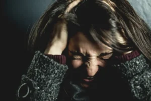 A person with long dark hair holds their head in both hands, eyes closed tightly and face tense, appearing stressed or overwhelmed, against a dark background.