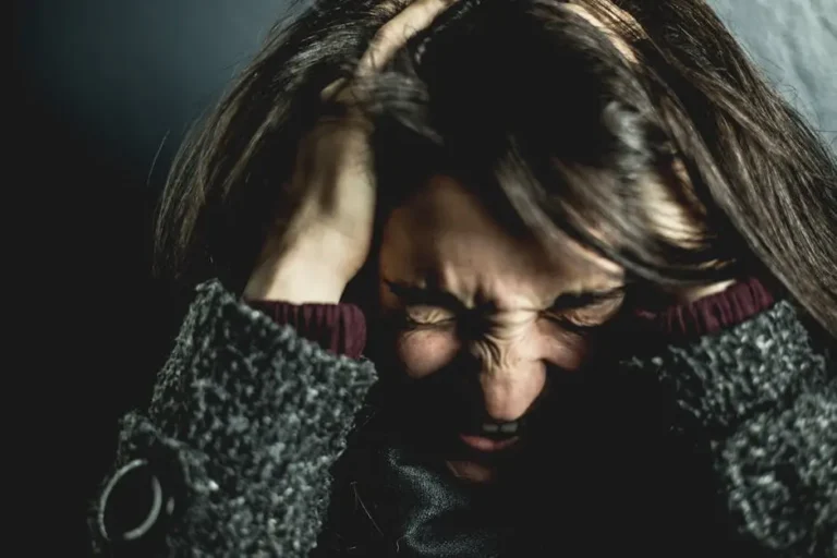 A person with long dark hair holds their head in both hands, eyes closed tightly and face tense, appearing stressed or overwhelmed, against a dark background.