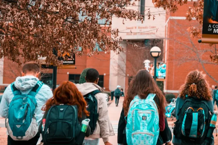 Five students with backpacks walk together on a college campus pathway, surrounded by trees and buildings, with leaves on the ground and some students in the background.