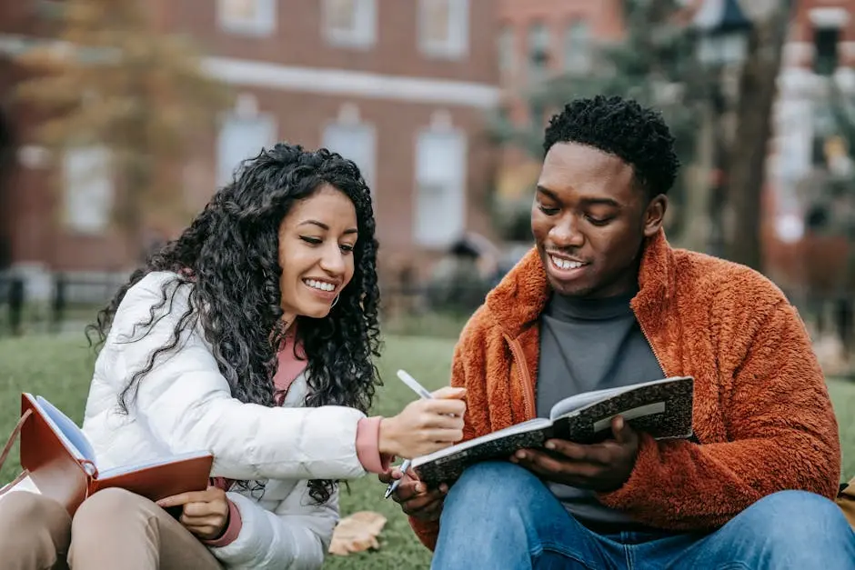 Two students sitting on grass, smiling and studying together; one is holding a notebook while the other writes in it. Trees and a brick building are visible in the background.