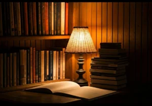 A cozy reading nook with a glowing table lamp, open book on a wooden desk, stacked books beside it, and shelves filled with neatly arranged books against a wood-paneled wall.