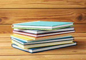 A stack of colorful notebooks with spiral and hardcover bindings sits on a wooden surface against a wooden background.