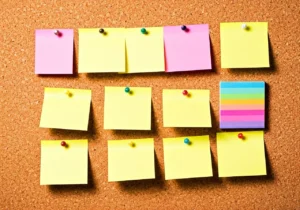 A corkboard with twelve yellow sticky notes, four pink sticky notes, and one stack of multicolored sticky notes, all pinned with colorful pushpins in three rows. The sticky notes are blank.