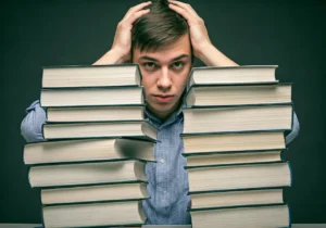 A young man sits behind two tall stacks of books, holding his head with both hands and looking stressed or overwhelmed.