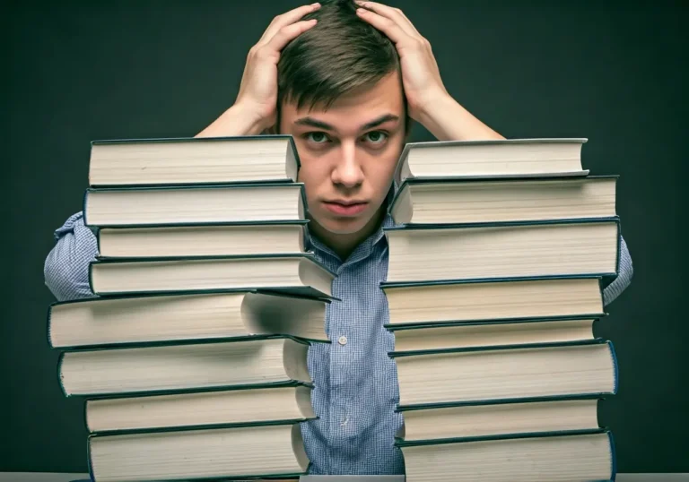 A young man sits behind two tall stacks of books, holding his head with both hands and looking stressed or overwhelmed.