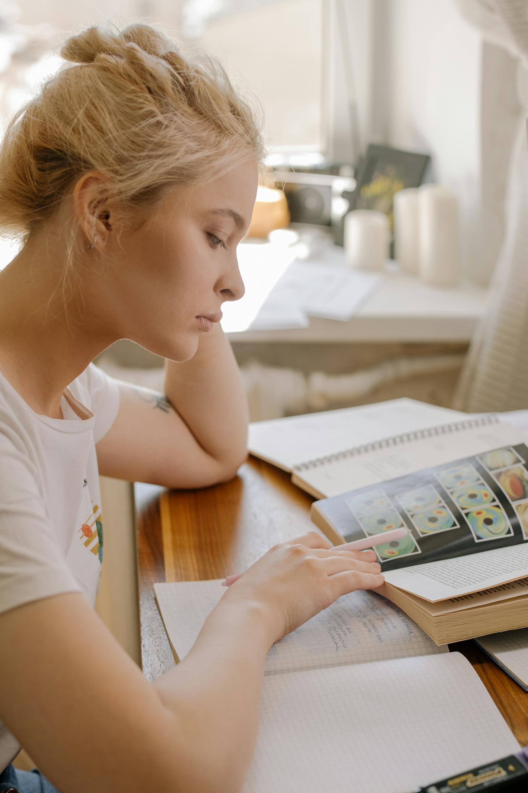 Focused young woman studying at home by the window with books open, illustrating a learning environment.