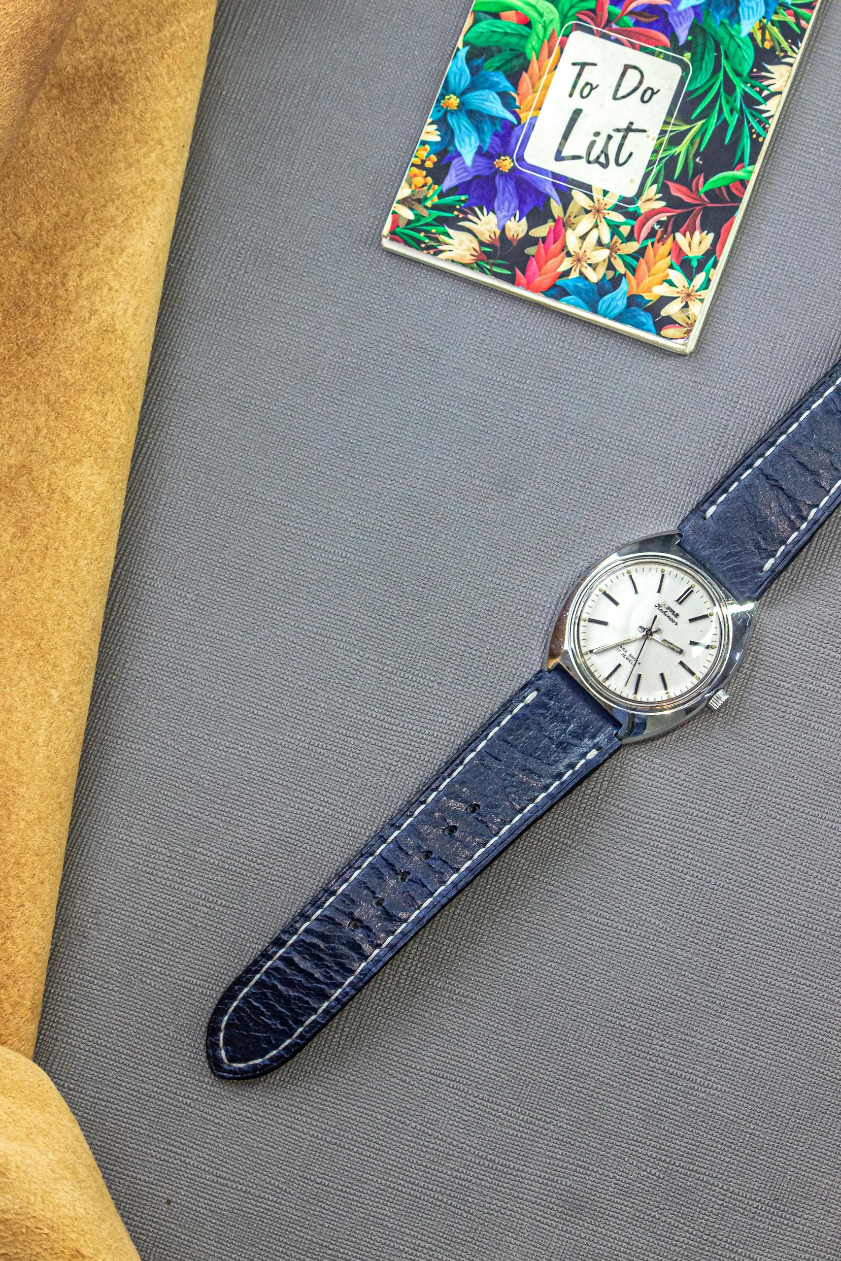 A vintage watch with a blue leather strap on a textured grey surface, featuring a colorful to-do list.