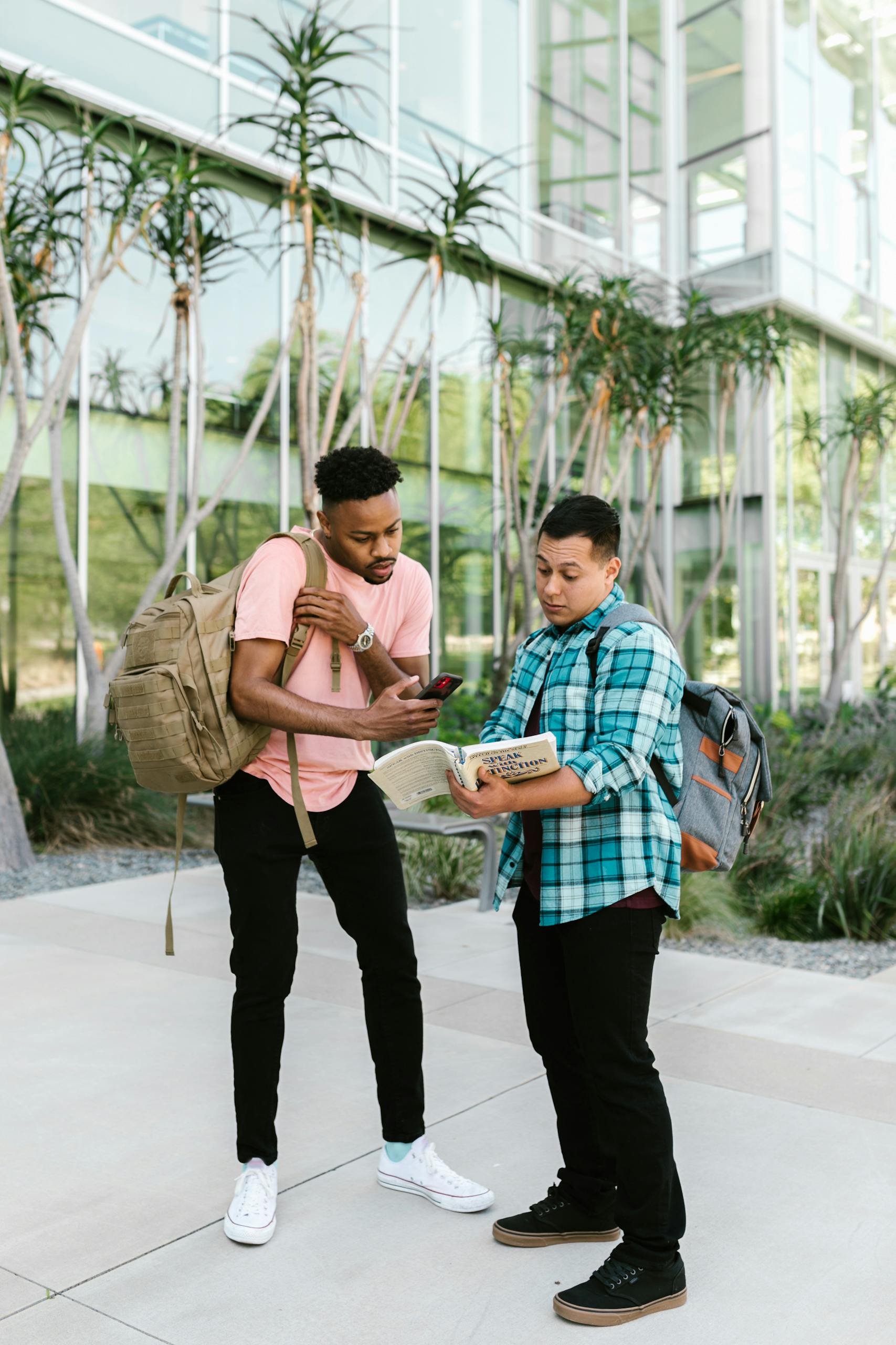 Two students with backpacks discuss study material on a sunny campus day, standing outdoors.