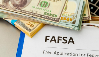 A stack of US dollar bills rests on top of a FAFSA form labeled Free Application for Federal Student Aid, with part of a laptop keyboard visible below.