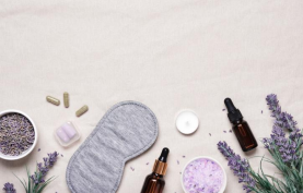 A gray sleep mask, lavender sprigs, bottles with droppers, a candle, capsules, and small bowls with lavender and bath salts are arranged on a light fabric background.