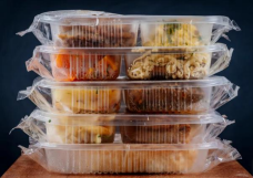 A stack of five clear plastic food containers filled with assorted cooked meals, placed on a wooden surface with a dark background.