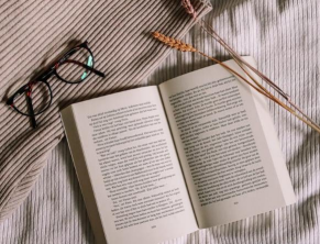 Open book on a bed beside a pair of black glasses and two dried wheat stalks, with a ribbed beige blanket partially visible underneath.